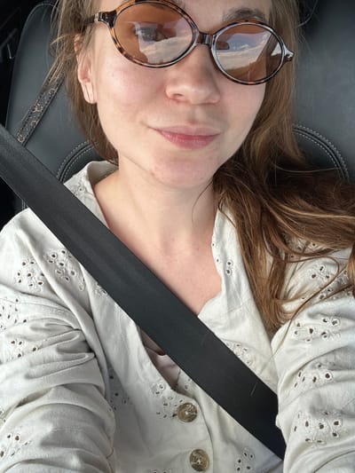 The image shows a close-up, selfie-style shot of a woman wearing tortoiseshell sunglasses and a white eyelet shirt. She is seated in a car, with a black seatbelt diagonally crossing her chest. The background includes the car's dark interior, specifically the headrest and stitching of the seat. The woman's hair is brown and appears to be pulled back loosely. Her expression is relaxed, with a slight smile. The reflection in her sunglasses shows a cloudy sky and what appears to be a landscape. The image does not provide any information or context to place it in the city of Pervomaiskoe, Russia.
