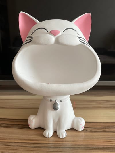 A white ceramic cat-shaped trinket holder sits on a wooden surface. The cat has pink inner ears, pink paw pads, black whiskers, and a large open mouth. It is wearing a silver tag around its neck. The background is dark and out of focus. The image does not contain any information about the city of Nefteyugansk, Russia.