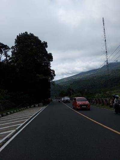 The image depicts a two-lane asphalt road in the city of Phoxay, Laos, under an overcast, gray sky. The road is marked with a solid yellow center line separating traffic, and a solid white line with diagonal white stripes delineates the left shoulder.

Vehicles observed include a red minivan with a visible front license plate, traveling towards the viewer in the right lane. Behind it, on the far right, a partially obscured motorcycle with a helmeted rider is present. Further down the road, multiple light-colored cars, including white sedans, are visible in both lanes, along with another motorcycle and rider.

To the left of the road, a dense forest of dark green trees rises sharply, bordering the road. A low stone retaining wall with black and white diagonal striped markings runs along the base of this forested area. To the right of the road, steep, forested hills and mountains ascend into the background. A tall, slender metal lattice tower, supporting multiple power lines, stands prominently on the right side of the scene, against the backdrop of the hills and cloudy sky. Utility poles are also visible further down the road on the right.