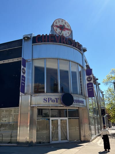 The image captures the exterior of a two-story building in Rochester, United States, with a distinctive architectural style. The building, identified as "SPOT Coffee," features a prominent "CHEVROLET SUPER SERVICE" sign with a clock above it. The clock shows an arrow through the Chevrolet logo. The façade is primarily composed of corrugated metal and glass, with purple banners displaying the "SPOT YOUR NEIGHBORHOOD CAFE" logo. The entrance doors are centrally located and appear to be papered-over, suggesting the business is closed or undergoing renovations.
A woman dressed in a white shirt and black pants is walking along the sidewalk in front of the building. A blue banner reads "EDEN COFFEE AND CO." A few trees are visible on the right side of the frame. The sky is clear and blue, indicating a sunny day.