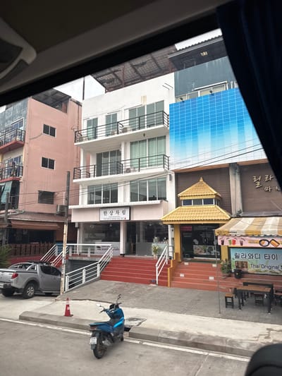 The image depicts a street scene in Bang Phra Subdistrict, Thailand, with a focus on several multi-story buildings. The foreground shows a blue motorcycle parked on the side of the road, with a red traffic cone nearby. A silver pickup truck is parked on the left, facing away from the camera.

The buildings are varied in style and color. On the left is a tall, pale pink building with multiple balconies and windows. Adjacent to it is a white building with several floors, also featuring balconies and numerous windows. One section of this white building is covered by a large, blue, grid-patterned advertisement. To the right of the white building is a brown building with a distinctive golden, multi-tiered roof structure above its entrance, which appears to be a restaurant or shop. This building also has an awning with yellow and white stripes. Signs in Korean and Thai are visible on the buildings, suggesting a mix of businesses.

The street itself is paved, with a sidewalk separating the road from the buildings. The environment appears to be a commercial or mixed-use urban area, with evidence of shops and possibly residential units. The sky is not clearly visible, but the overall lighting suggests it is daytime.