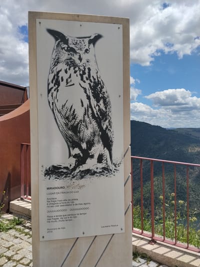 The image shows a vertical sign located in Alijó, Portugal, at the Miradouro da Fraga do Újo (Újo Rock Overlook). The sign is white with a black stenciled image of an owl and text in Portuguese. The text includes the name of the overlook, a short poem by Laureano Ribatua, and the name of the municipality, Alijó, and the year 2019. The poem describes the appearance of the owl (Újo) at night, with its hooting, and mentions the legend associated with the rocks, the moon, and the owl. The background includes a glimpse of the landscape, including mountains, green trees, and a sky with clouds. A red metal railing and a staircase are visible near the sign, along with some vegetation growing in the immediate foreground.