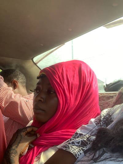 The image depicts a woman wearing a red headscarf. She is looking down with a somber expression. A child with short hair is leaning against her. The woman has intricate henna on her hand and is adjusting her scarf. Another person, wearing a white patterned shirt, is visible in the upper right. The setting appears to be inside a vehicle, indicated by the interior ceiling and window frame. The image was taken in the city of Jiru, Nigeria.