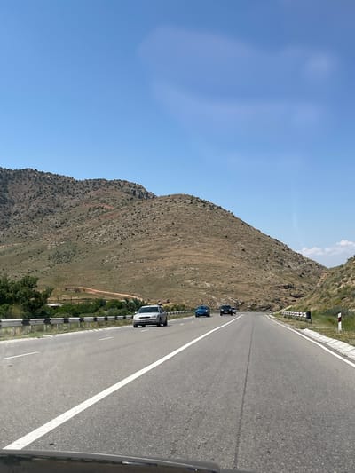 The image displays a mountain road scene in Zangakatun, Armenia. The primary subjects are the road, a steep, arid mountain slope, and several cars traveling on the road. The scene is set during the day, with clear blue skies, suggesting fair weather.
The road is asphalt, with a white line marking the center and edge. It curves gently to the right as it ascends. Several vehicles are visible on the road, including a dark-colored car, a blue car, and a white SUV. The cars appear to be moving along the road, indicating travel or transit.
The mountain slope on the left of the road is characterized by dry, sparse vegetation and rocky outcrops. The terrain is rugged and undeveloped. On the right side of the road, there is a guardrail with alternating black and white reflective markers, commonly found along highways and mountain passes. Beyond the guardrail, there are some trees and glimpses of distant structures.
A notable detail is the presence of traffic signs or markers on the side of the road, including striped poles with red and white markings. The perspective of the image suggests it was taken from a moving vehicle, likely the front passenger seat or dashboard, looking forward and slightly to the left.
There is no visible text in the image, aside from what might be license plates on the vehicles, which are too small to be legible. The overall impression is one of a scenic drive through a mountainous landscape.
