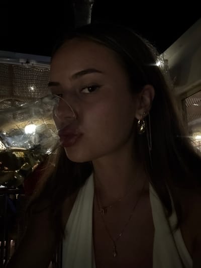 A female subject is captured in a close-up portrait, positioned in a dimly lit, possibly indoor or semi-outdoor, evening environment. She has long, dark hair and wears a light-colored halter-neck garment, adorned with multiple delicate necklaces, one featuring a butterfly charm and another an elongated pendant. Her right ear displays multiple piercings, including a gold-toned hoop earring and a slender chain earring. She is looking directly at the camera with her lips pursed, and a transparent, stemmed glass is held in front of her face, partially obscuring her nose and mouth while reflecting ambient light. The background consists of dark, indistinct shapes and a decorative screen or netting illuminated by small, warm-toned lights, characteristic of a bar or restaurant setting. The precise location within the city of Heraklion, Greece, is not discernible from the visual information presented.