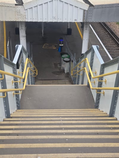 The image shows a view looking down a set of outdoor stairs with yellow and dark grey striped steps. The stairs are flanked by a white metal railing with yellow handrails. Beyond the stairs, an overhead walkway with a white corrugated roof supported by grey metal beams is visible. Below the walkway, a paved area with a yellow line marking and a blue recycling bin are present. Further in the background, a train track runs alongside a platform. The location is identified as Elmbridge, United Kingdom.
