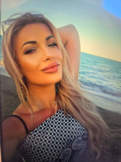 A fair-skinned woman with blonde hair is lying down on a beach. The ocean is visible in the background. The woman is wearing a black and white patterned top.