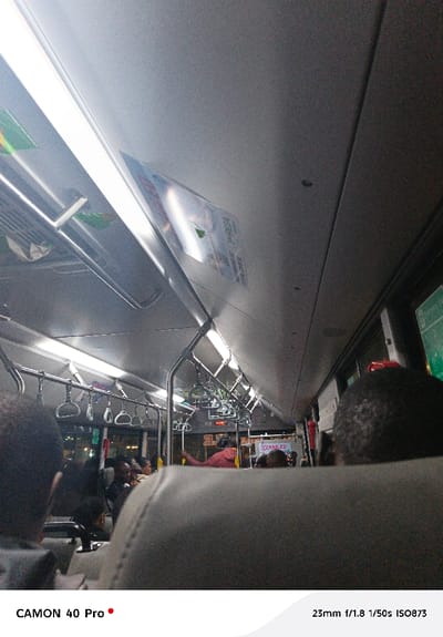 Inside a brightly lit bus, rows of silver handrails and hanging grips line the ceiling above seated passengers. At the back, a digital display glows. The view is from behind a high-backed seat, focusing on the bus's interior. A poster is affixed to the ceiling panel. The lighting consists of long, fluorescent lamps. People are seen occupying many of the seats. The atmosphere inside suggests a routine commute.