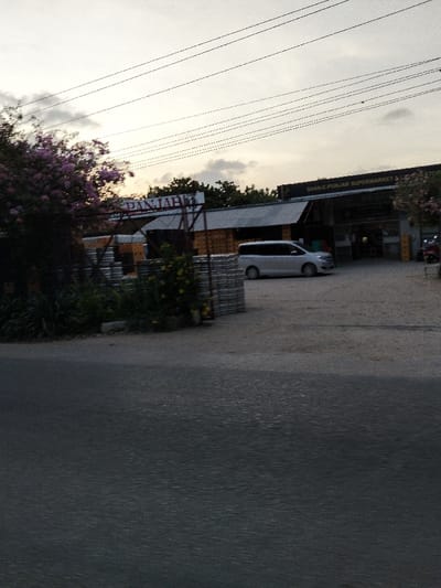 The image displays an outdoor scene in Gombato Bongwe ward, Kenya, during what appears to be twilight conditions. In the foreground, a dark asphalt road occupies the lower portion of the frame. Beyond the road, a light-colored gravel or dirt area serves as a parking lot leading to commercial establishments. On the left, a structure with wooden walls and a corrugated metal roof features a sign partially visible, displaying the text "DANJAB". In front of this structure, stacks of grey concrete blocks are arranged, flanked by green foliage including a tree with purple blossoms and a plant with yellow flowers. To the right, a building identified by a prominent sign as "SHAN-E-PUNJAB SUPERMARKET & LIQUOR STORE" is visible, also featuring a corrugated metal roof and an illuminated entrance. A light-colored minivan is parked in the gravel area between the two buildings. A red scooter or small motorcycle is parked near the entrance of the supermarket. Overhead, multiple dark power lines traverse the overcast sky horizontally. No individuals are discernible in the scene.