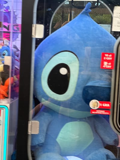 The image shows a large blue plush toy, resembling the character Stitch from Disney's Lilo & Stitch, inside a glass display case, likely a prize machine. The toy is predominantly blue with lighter blue accents on its belly and around its eyes. Its large, oval eyes are black with a white pupil. Red stickers are affixed to the glass in front of the toy. One sticker reads "10 zł 2 GRY" and another "10 zł 4 GRY", indicating pricing for games. A third sticker features a coin and the text "1 GRA". Below these, another red sticker has white text that is partially obscured but appears to say "AUTOMAT NIE WYMAGA". The display case has a metal hinge visible on the left side. In the background, through the glass and to the left, other plush toys are visible, suggesting a selection of prizes. The overall environment appears to be an indoor amusement area or arcade, possibly within a shopping center or similar establishment in Wolsztyn, Poland. The lighting is bright, reflecting off the glass.