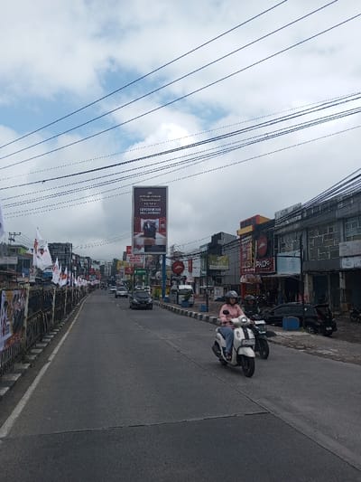 This image displays a bustling urban street scene under an overcast sky, with numerous black overhead power lines crisscrossing the frame. A multi-lane asphalt road stretches into the distance, with vehicle traffic moving in both directions.
In the foreground on the right side of the road, a person wearing a pink long-sleeved shirt, blue jeans, and a grey full-face helmet is operating a white scooter. The scooter's license plate, prominently visible, reads "D 3470 UZG". Directly behind and slightly to the left of the scooter, another individual on a black motorcycle, also wearing a helmet, is visible.
The right side of the street is lined with commercial buildings, many of which are storefronts. Visible signage includes "PHD Pizza Hut Delivery," the "Pizza Hut" logo, "DABLE STORE," and "MAJA BARBERSHOP." Several motorcycles are parked along the curb in front of these establishments. Blue, rectangular bollards or planters are spaced along the sidewalk edge.
On the left side of the road, a black metal fence or barrier extends into the distance, adorned with numerous white flags featuring red emblems, suggesting a political or commercial campaign. Further down the street, a prominent vertical billboard displays an advertisement with text, including "PT. PURNAMA NIAGA ABADI," and an image of a person seated. Several cars, including a black SUV and a white van, are visible in the background traffic.
While the prompt specified the location as Thotagamuwa, Sri Lanka, the visual evidence, specifically the scooter's Indonesian license plate format (D 3470 UZG) and the names of businesses like "PT. PURNAMA NIAGA ABADI," strongly indicates this scene is located in an Indonesian city.