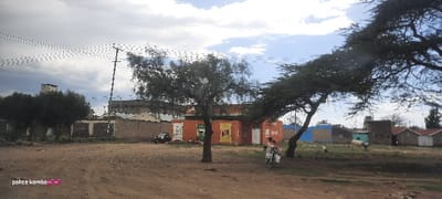 A street scene shows a dirt road with various structures visible. An orange building with posters is prominent, along with smaller shacks and houses. Trees provide shade, while electrical poles and wires traverse the sky. A motorcycle is seen on the right, and a car is parked near the orange building. The sky is partly cloudy, with a mix of gray and blue.