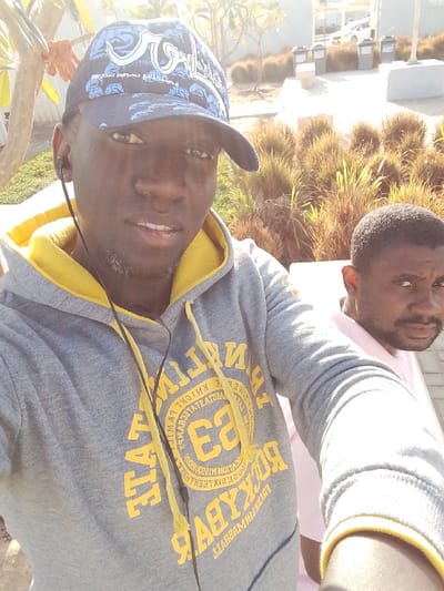 An outdoor selfie-style photograph depicts two males in a landscaped area within النعيلة, Saudi Arabia, under bright daylight conditions. The primary subject, a dark-skinned male, faces the camera directly, exhibiting a neutral expression with a slightly open mouth. He wears a blue baseball cap adorned with white patterns and a grey hoodie featuring a yellow inner lining and yellow text, including "FRANKLIN" and "83" within a circular emblem design. Black wired earphones are visible around his neck and entering his left ear. To the right and slightly behind the primary subject, a second dark-skinned male is partially visible, oriented towards the right side of the frame. He has short dark hair and a beard, and appears to be wearing a light-colored shirt. The background features rows of ornamental grasses, a paved walkway, two grey trash receptacles, a light-colored bench, and indistinct buildings with a white vehicle in the far distance.