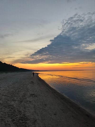 A lone figure walks along a sandy beach at sunset in Apšuciems, Latvia. The sky is a gradient of grey clouds above a vibrant orange and yellow horizon, reflecting on the calm water. A dark tree line borders the left side of the beach. The sand is uneven and marked with footprints.