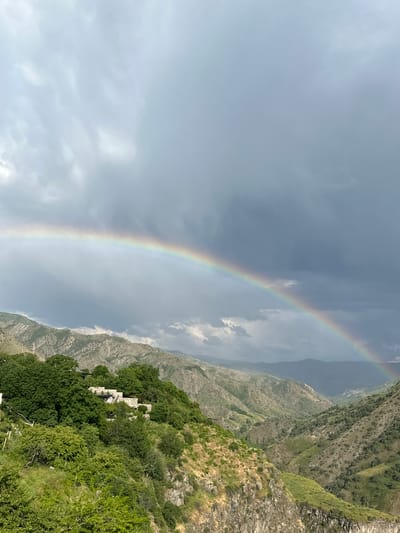The image depicts a dramatic landscape in Garni, Armenia, featuring a vibrant rainbow arcing across a cloudy sky over a rugged, mountainous terrain. The foreground is dominated by lush green trees and dense foliage clinging to steep slopes, with hints of dry grass and rock formations. A series of stone structures, possibly buildings or ruins, are nestled amongst the trees on the hillside. The middle ground reveals the undulating contours of the mountains, characterized by a patchwork of green vegetation and rocky outcrops, with deep valleys carved between them. The sky above is a mix of dark, heavy clouds and lighter patches, with the rainbow serving as a striking contrast. The overall scene suggests a post-rain atmosphere in a natural, somewhat wild environment. There are no people visible in the image.