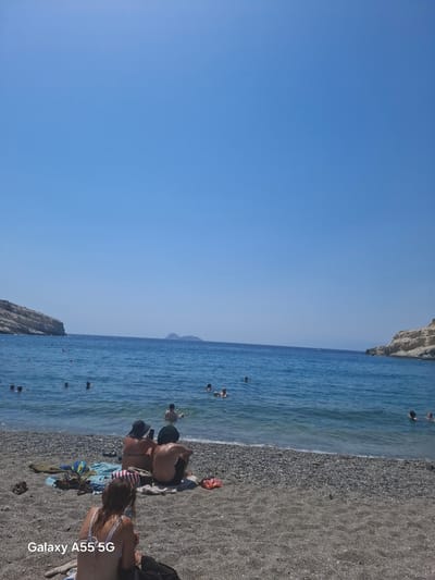 This media file depicts a sunny day at a pebble beach in Tybakio Municipal Unit, Greece. The scene is divided into two main areas: a clear blue sky occupying the left half of the frame, and the ocean and beach on the right. The water is calm and a deep blue, with several people swimming and wading. The beach itself is covered in dark grey pebbles, and some beach towels and bags are scattered on the sand.

In the foreground, several people are sitting on the beach, facing the water. One person in a bikini is looking out at the sea, and a couple is sitting together. Further out in the water, more individuals are visible swimming. The overall atmosphere appears relaxed and leisurely, suggesting a typical summer day at the beach.

To the left of the water, a rocky cliff face rises, adding to the natural beauty of the location. In the distance, a faint mountain range can be seen on the horizon. The bright, clear sky indicates it is daytime, likely mid-morning or afternoon, under fair weather conditions.

The bottom right corner of the image has white text overlay which reads "Galaxy A55 5G".