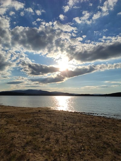 The image depicts a tranquil waterscape with a sandy shore in the foreground. The sun is partially obscured by scattered clouds, casting a shimmering reflection on the water's surface. In the distance, a chain of dark hills stretches across the horizon, silhouetted against the bright sky. The beach is composed of sand and small stones, with sparse patches of dry grass. The overall scene conveys a sense of calm and natural beauty. The location is a lake in the vicinity of Radslav, Czechia, known for its scenic natural surroundings. There are no people or specific objects of interest in the scene, aside from the natural elements.