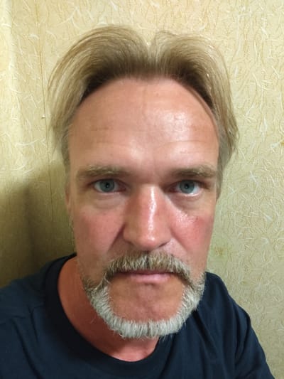 The image is a close-up, head-and-shoulders selfie of a middle-aged Caucasian male with light brown hair, a goatee and mustache, and blue eyes. He is wearing a dark blue t-shirt. The background is a textured yellow wallpaper. There is no visible indication of the location in the city of Rezekne, Latvia.