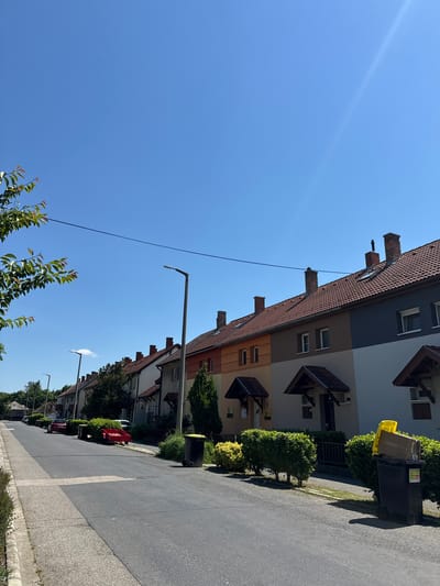 The image depicts a residential street in Siófok, Hungary, characterized by attached houses with sloping red tile roofs, chimneys, and projecting decorative window coverings. The buildings are painted in a gradient of gray, brown, and orange hues. The street has a gray asphalt surface with a lighter concrete curb. Green bushes and trees line the street. Utility poles and wires are visible against a clear blue sky. Garbage bins are positioned near the houses. Parked cars are visible further down the street.