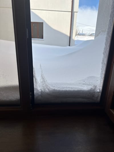 The scene viewed through a window reveals an immense amount of snow, nearly obscuring the entire view. The snow is piled high outside, reaching the top of the window frame, with a visible curvature indicating depth. A building partially visible in the background, juxtaposes the snow. The window frame itself, a dark color, provides a sharp contrast to the bright, white expanse of snow. The inside of the room is dimly lit, with a dark wooden wall on the right.
