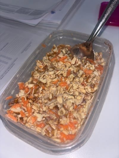 A plastic container of food sits on a white surface, partially obscuring stacks of papers. The food appears to be a mixture of oats, chopped carrots, and nuts in a liquid, possibly milk or yogurt. A spoon rests in the container, presumably for eating. The environment suggests an indoor setting, likely a desk or table due to the presence of paperwork. The location of Buenavista, Mexico, is not discernible from the image.