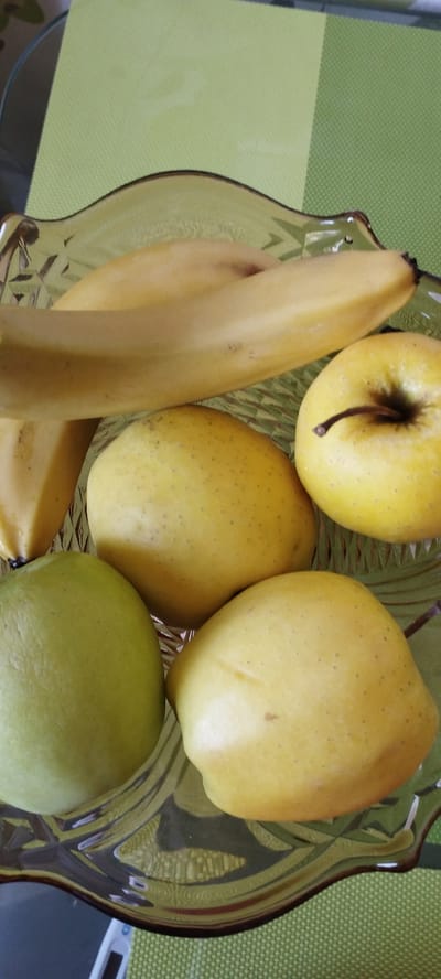 A fruit bowl sits on a green placemat. The bowl is made of clear, patterned glass and contains a banana, three yellow apples, and one green apple. The banana is positioned horizontally across the top of the bowl, and the apples are arranged below it. The scene appears to be indoors, possibly in a kitchen or dining area. The presence of fruit suggests a domestic setting. There is no indication of the specific location within Skidzieĺ, Belarus, beyond the general fact that it is in that city. The image is taken from a slightly elevated angle.