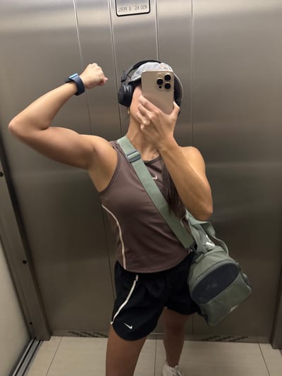 In an elevator, a person wearing a brown Nike tank top, black Nike shorts, and white sneakers poses for a selfie. They have a black smartwatch on their left wrist, black headphones on, and are holding a gold iPhone in their right hand to take the photo. A light green gym bag with navy blue accents hangs from their left shoulder. The elevator's brushed metal walls are visible, along with a sign at the top displaying text in reverse. The floor is tiled.