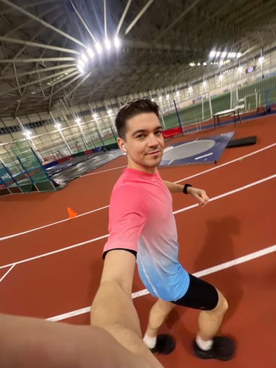 A man stands on a red indoor track, facing the camera. He wears a pink and blue gradient shirt, black shorts, and black shoes with white socks. He has short dark hair, a white earbud in one ear, and a black watch on his left wrist. The track is marked with white lines. A large metal structure is visible in the background, along with stadium lights. The photo appears to be a selfie taken indoors.