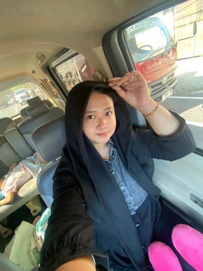 The image is a selfie taken from inside a vehicle, likely a van, traveling on a street in Ngrame, Indonesia. A woman wearing a black hijab and a denim shirt is in the foreground, looking at the camera. Her right hand is near her head, adjusting her hijab, and she is wearing a bracelet. In the background, through the side window, a red truck with the text "ANGKUT" is visible, along with other vehicles and buildings. To the left of the woman, a child is seen sleeping in a seat. The interior of the vehicle shows multiple rows of grey seats. The overall lighting suggests it is daytime, possibly late afternoon due to the warm tones.