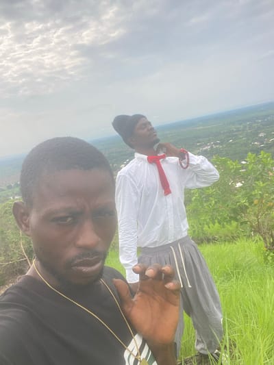 Two individuals are positioned on a grassy hilltop overlooking a sprawling landscape in Amaigbo, Nigeria. The person in the foreground, facing the camera, is wearing a black t-shirt and a gold chain necklace. Their left hand is raised, fingers splayed, in a gesture that obscures a portion of their face and chest. The second individual, in the background, is wearing a black beanie, a white long-sleeved shirt, and grey sweatpants. A red scarf is tied loosely around their neck, and they have bracelets on their right wrist. This individual's head is tilted back, with their eyes closed or looking upwards. The environment is characterized by lush green grass and foliage, with a distant view of the town of Amaigbo, Nigeria, its buildings and structures visible amidst the greenery. The sky above is overcast with scattered clouds.