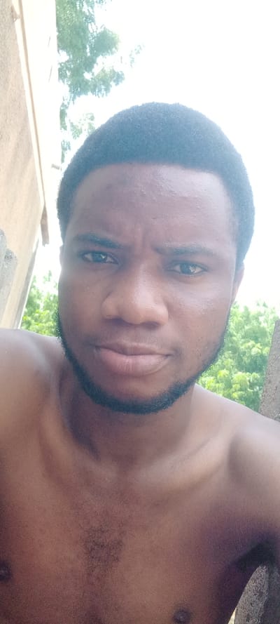 A close-up, head-and-shoulders portrait depicts a male individual, appearing to be a young adult, with dark skin, short black hair, and a neatly trimmed beard and mustache. He is shirtless, with the upper chest and shoulders visible. The subject is centered and looks directly into the camera with a neutral expression. The background indicates an outdoor setting with bright, diffused light. To the left, a light-colored building wall is partially visible. Behind the subject, green foliage from trees is present against a bright, possibly overcast, sky. A dark, textured surface, possibly concrete or stone, is partially visible in the lower right foreground. The specific city location of Mairi, Nigeria, cannot be determined from the visual information presented in the image.