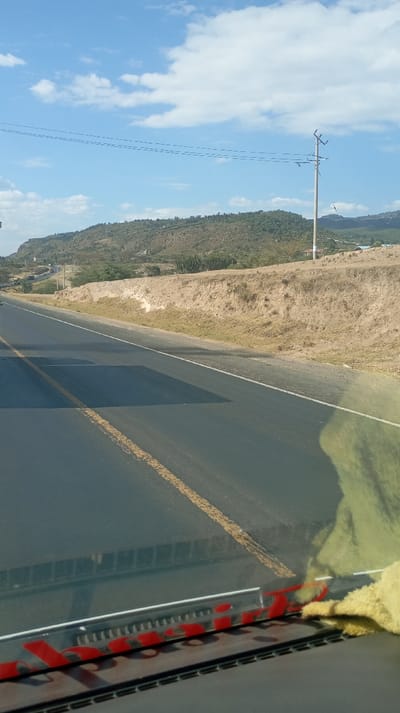 A road stretches into the distance, marked by yellow lines. Beyond the road, a low hill rises, dotted with vegetation and topped with a ridge. A utility pole stands to the right of the hill. Above, a blue sky is scattered with white clouds. The image appears to be taken from inside a vehicle, as part of the dashboard is visible at the bottom.