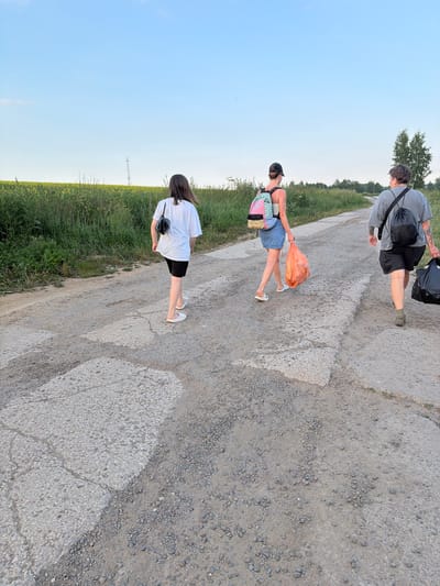 The image shows three people walking on a dirt road, bordered by a grassy field and a clear blue sky. The time of day appears to be late afternoon or early evening due to the soft lighting and long shadows.
The person in the foreground is a woman wearing a white t-shirt and black shorts, carrying a black bag slung across her chest. She is walking away from the camera, her feet clad in white shoes.
Behind her, another woman is walking, also away from the camera. She is wearing a denim skirt and a pink top, and she is carrying a colorful backpack. In her right hand, she holds a bright orange plastic bag filled with objects, possibly groceries or trash. She is wearing white sandals.
Further up the road, a third person, who appears to be a man, is walking towards the camera. He is wearing a grey t-shirt, black shorts, and dark shoes. He has a black backpack on his left shoulder and is carrying a black trash bag in his right hand. His arms are covered in tattoos.
The road itself is unpaved and appears to be made of gravel and dirt, with some larger stones and concrete patches. The field beside the road is lush and green, with some tall grass and scattered wildflowers. In the distance, on the horizon, are a few trees and what looks like a telecommunications tower. The overall atmosphere is casual, suggesting a leisurely walk or perhaps a journey. No text is visible in the image.
