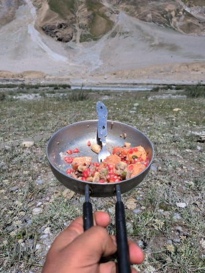 A hand holds a metal cooking pot containing a mixture of food. The pot has two black handles, one of which is being held by the hand. A silver fork rests inside the pot, embedded in the food. The food appears to be a reddish-orange color with visible pomegranate seeds and other chunky ingredients. The pot is positioned in front of a natural landscape. In the background, a body of water flows between rocky, barren mountains. The ground in the foreground is covered with dry grass, small stones, and sparse vegetation. The sky is clear and bright, suggesting a daytime scene. The overall impression is of an outdoor meal prepared in a rustic, remote location, possibly in the mountainous region of Matayeen, India.