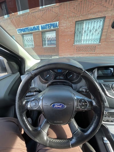 This is a close-up, interior shot of a car, likely a Ford, viewed from the driver's perspective. The steering wheel, dashboard with gauges and buttons, and parts of the car's interior are the primary focus. Outside the car, a brick building with windows is visible, suggesting an urban or suburban setting. The windows have decorative metal grates. Text on the building reads "СТРОИТЕЛЬНЫЕ МАТЕРИАЛ", which translates to "BUILDING MATERIALS". The car's dashboard displays the time as 17:58 and the date as 01.04.2014, indicating it's late afternoon on April 1st, 2014. The car's odometer shows "156484км". The lighting suggests it's daytime and sunny, with the sun casting shadows. There are no people visible in the image. The overall impression is that of a parked car, with the driver taking a picture of the view outside or the car's interior.