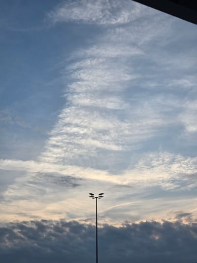 The image is a vertical, medium-shot photograph of a twilight sky with scattered clouds and a lamppost in the foreground. The sky transitions from a light blue near the horizon to a darker blue at the top. Wispy, white cirrus clouds are visible across the upper portion of the sky, with a more dense, dark gray layer of stratocumulus clouds occupying the lower third. A tall, thin lamppost with two heads is silhouetted against the sky in the right half of the frame. The top edge of the image is partially obscured by a dark, horizontal bar, suggesting it might be taken from indoors or under an overhang. The image does not provide any specific visual cues to pinpoint its location within Ansbach, Germany.