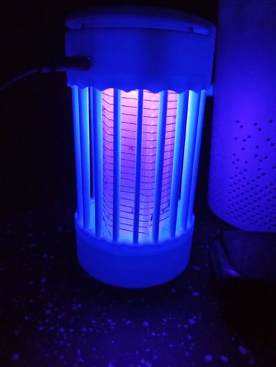 An electric insect killer is displayed against a dark background. The device is cylindrical with a white plastic casing and a grid of blue rods. Inside the rods, a light source emits a purple glow. The device is connected to a black cable. To the right of the insect killer, a taller, white, perforated cylindrical object is partially visible. The surface beneath the objects is dark and speckled with small white and purple flecks. The scene is dimly lit, with the blue and purple light from the insect killer dominating the illumination.
