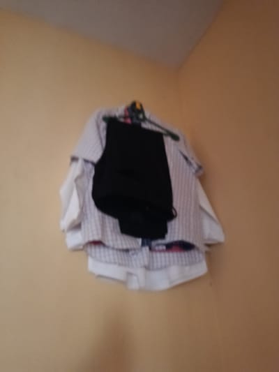 In Ukpor, Nigeria, a black pair of pants is draped over a white and red checkered shirt, which is then hung on a green clothes hanger. The clothes are displayed against a light yellow wall, with the corner of the room visible in the background. The image is slightly out of focus, obscuring finer details.