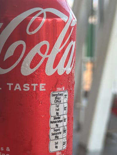 An extreme close-up, slightly tilted, side-view of a cold, condensation-covered red Coca-Cola can. The iconic white Coca-Cola logo is prominently displayed across the can's surface. To the right of the logo, a nutrition facts label is visible, detailing energy content, fat, saturated fat, sugars, and salt. The background is blurred, suggesting an outdoor environment with diffused natural light and hints of muted green and beige structures. There are no people or animals present in the image, and no specific indicators place the scene in Constanța, Romania.