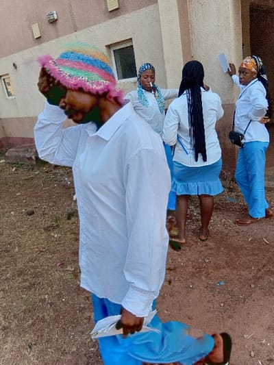 Four individuals are positioned outside a building. Three appear to be uniformed, wearing white shirts and blue skirts or pants, with head coverings on two of them. The fourth person is closer to the viewer, dressed in a white shirt and blue pants, wearing a colorful knitted hat. All are standing on a dusty ground area adjacent to a building with light-colored walls and visible windows.