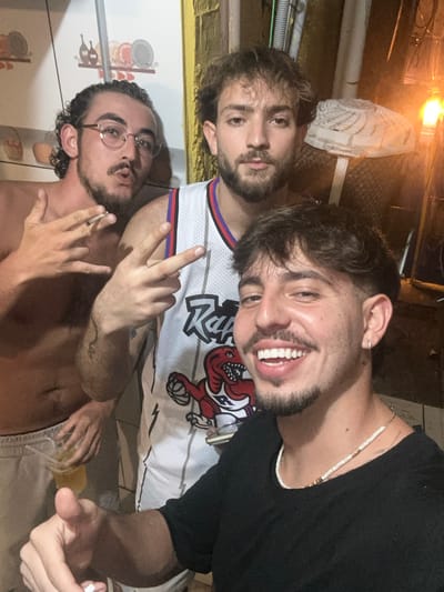 Three young men are posing for a photo. One shirtless man wears glasses and holds a cigarette. Another wears a white jersey with a dinosaur logo. He's flashing a hand sign and smiling broadly. The third man is in the background, partially visible behind the other two. The image is rotated. The background is a mix of indoor and outdoor elements, including a fan and a tiled wall.