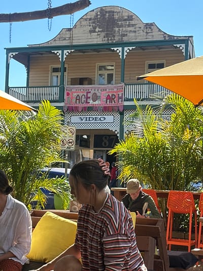 The image captures a sunny outdoor scene in Bellingen, Australia, likely a cafe or restaurant. In the background, a two-story wooden building with a distinctive curved roof is visible. A banner that reads "PEACE ON EARTH" hangs above the entrance, which also has a sign that says "VIDEO." The building appears to be at address number 87. In the foreground, several people are seated at tables. A young woman in a striped shirt is in the process of being seated, while another woman in a white shirt and patterned pants is already seated. An older man wearing a cap and vest is seated at a table, reading. Orange chairs are visible, suggesting outdoor seating. Lush green palm trees provide shade and a tropical feel to the environment. The overall atmosphere is relaxed and casual.