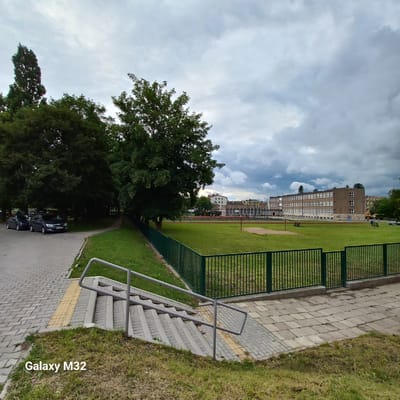 The image depicts an outdoor scene at what appears to be a school or sports complex in Olsztyn, Poland. In the foreground, there are concrete stairs with metal handrails leading up from a grassy area to a paved walkway. The walkway is constructed of interlocking gray and yellow paving stones. To the left of the walkway, several cars are parked on a similar paved surface, with mature trees lining the area.
The middle ground features a large, fenced-off grassy field, likely a sports field, with a red running track visible in the distance. Beyond the field, a multi-story brick building with numerous windows stands prominently, suggesting it is a school or public institution. Other smaller buildings and trees are visible in the background, under a cloudy sky. The overall impression is one of an urban educational or recreational space on a slightly overcast day. The watermark "Galaxy M32" is present in the lower left corner of the image.