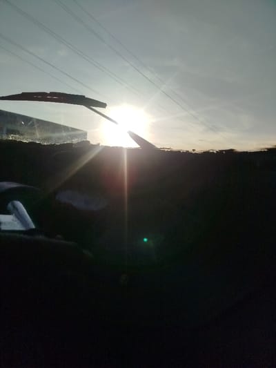 The image displays an outdoor scene in Keekonyokie ward, Kenya, captured from the interior perspective of a vehicle looking forward through its windshield. The dominant feature is a bright, low-lying sun, positioned slightly to the right of the frame's vertical center, casting significant lens flare that radiates across the lower-left and central portions of the view. The sky above is a pale blue, with several parallel overhead power lines stretching horizontally across the upper third of the frame. A black windshield wiper blade is visible across the upper left quadrant of the windshield. The top edge of the vehicle's dashboard or cowl is silhouetted prominently across the lower portion of the windshield. The vehicle's interior below the windshield is largely underexposed and appears dark, with a distinct green lens flare visible in the lower right corner. No individuals or overt actions are discernible in the scene.