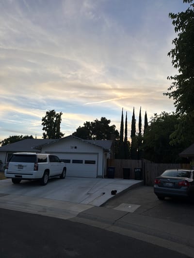 The image depicts a residential street in Citrus Heights, California, USA, under a cloudy sky with streaks of orange and yellow suggesting either sunrise or sunset. In the foreground, asphalt pavement leads to a driveway. A white GMC Yukon XL SUV is parked in the driveway, facing away from the camera, in front of a white garage with a sectional door. To the right of the Yukon, a dark-colored sedan is parked on another section of the driveway. A black cat is visible sitting on the concrete slope of the driveway, near a manhole cover. The house itself appears to be a single-story structure with a grey shingled roof. Behind the house, a wooden fence separates it from neighboring properties. Several tall, dark green, columnar trees stand in a line behind the fence, silhouetted against the sky. Trees and foliage are visible on the right side of the image, partially obscuring the view of the sky and neighboring houses. Two dark trash bins with the number "6301" stenciled in white are positioned near the fence.