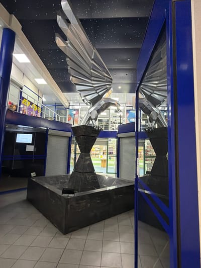 This media file showcases a modern, interior scene that appears to be a public space, possibly a museum, gallery, or exhibition hall. The dominant feature is a large, abstract metallic sculpture resembling a stylized bird with outstretched wings. The sculpture is intricately faceted, catching and reflecting light, giving it a dynamic and futuristic appearance. It is positioned against a dark, textured wall dotted with small, bright points of light, creating a "starry sky" effect.
The sculpture is framed by bold, blue architectural elements, suggesting a contemporary design aesthetic. Reflections in the sculpture and polished surfaces reveal glimpses of the surrounding environment, including shop windows or display areas stocked with various goods. These reflections also show more of the architectural features, such as roll-up shutters and illuminated panels.
The overall setting is indoors, with artificial lighting contributing to the ambiance. There are no people visible in the frame. The scene is devoid of any specific weather cues or time-of-day indicators, other than what is suggested by interior lighting. The presence of the metallic sculpture and the reflective surfaces hints at an artistic or informational exhibition.
The crops provide closer views of the metallic sculpture, the starry wall, and the architectural details, reinforcing the overall impression of a contemporary public space designed to display art or information in an engaging and visually striking manner. No visible text is clearly discernible in the original image or the provided crops that would offer further context about the specific location or exhibits.