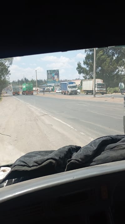 A view from inside a vehicle shows a sunny, multi-lane road. Three large trucks are traveling on the left side of the road, and two are parked or driving slowly on the right. Several other vehicles are also visible on the right side of the road. A billboard stands on the right, displaying an advertisement. Trees are scattered along the roadside. The interior of the vehicle is visible at the bottom of the frame.