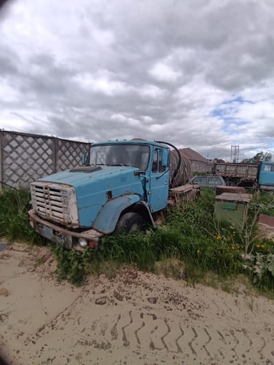 The image depicts a weathered, light blue truck partially submerged in tall grass and sand in an open area in Яльгелево, Russia. The truck's front end, including the grille and bumper, are visible, and the driver's side door is partially open. The vehicle appears to be abandoned or not in use. Other vehicles, including another truck with a flatbed, are visible in the background. A concrete block wall is visible behind the front truck. The sky is overcast with cloudy conditions. The ground is covered in a mix of sand, grass, and tire tracks.