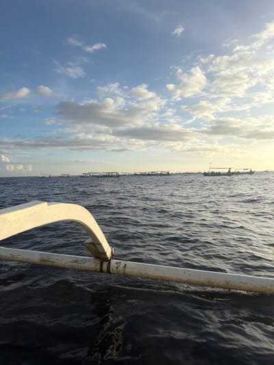 The scene depicts a marine environment off the coast of Pengastulan, Indonesia, viewed from the perspective of an outrigger vessel. In the immediate foreground, the weathered white outrigger structure of the observation boat, consisting of a curved arch and a horizontal pole secured by bindings, extends diagonally into the lower left portion of the frame over dark, rippled ocean water. The midground features a prominent horizontal line of approximately twenty-five traditional Indonesian outrigger boats, each equipped with canopies and occupied by multiple human figures, appearing clustered and relatively stationary on the water's surface. The background consists of a clear horizon line separating the ocean from a partially cloudy sky, which transitions from light blue at the top to a brighter, warmer hue on the right, indicative of a low sun angle. No specific individual actions or interactions are discernible among the distant figures; however, their collective presence on numerous vessels suggests a coordinated activity.