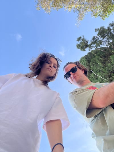 Two individuals, a male and a female, are positioned above the camera, gazing downward. The male, adorned with sunglasses, wears a light green shirt with a graphic. The female, with her own pair of sunglasses perched on her head, dons a white shirt. Trees and the blue sky serve as the backdrop. The location is Germasogeia, Cyprus.